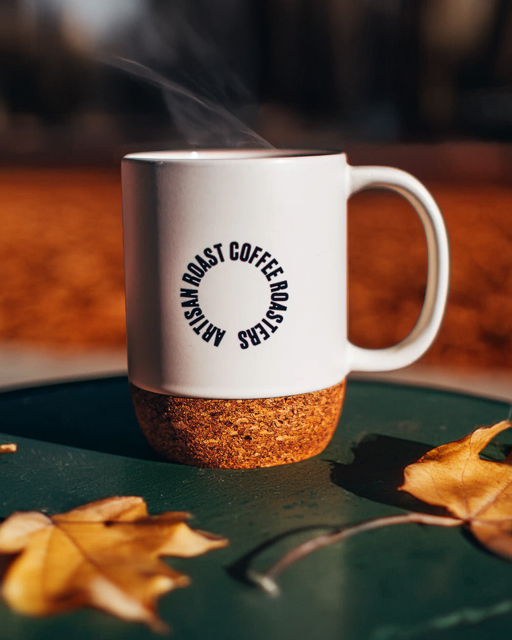 White mug with cork base on a green surface with autumn leaves, blurred background