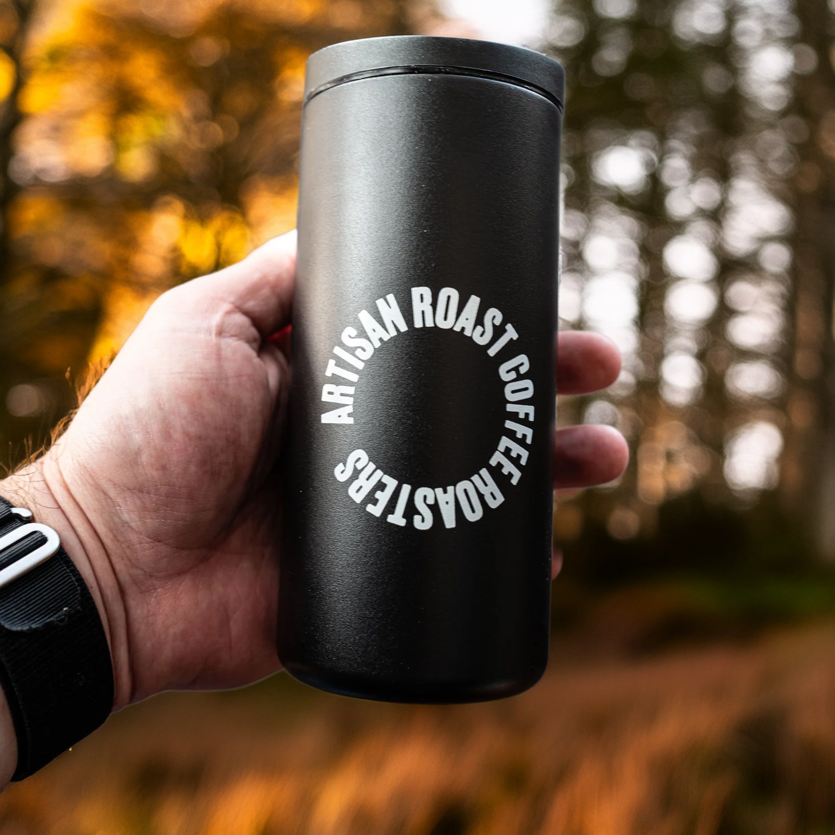 Black insulated coffee cup with 'Artisan Roast Coffee Roasters' logo held in a hand against a blurred forest background.